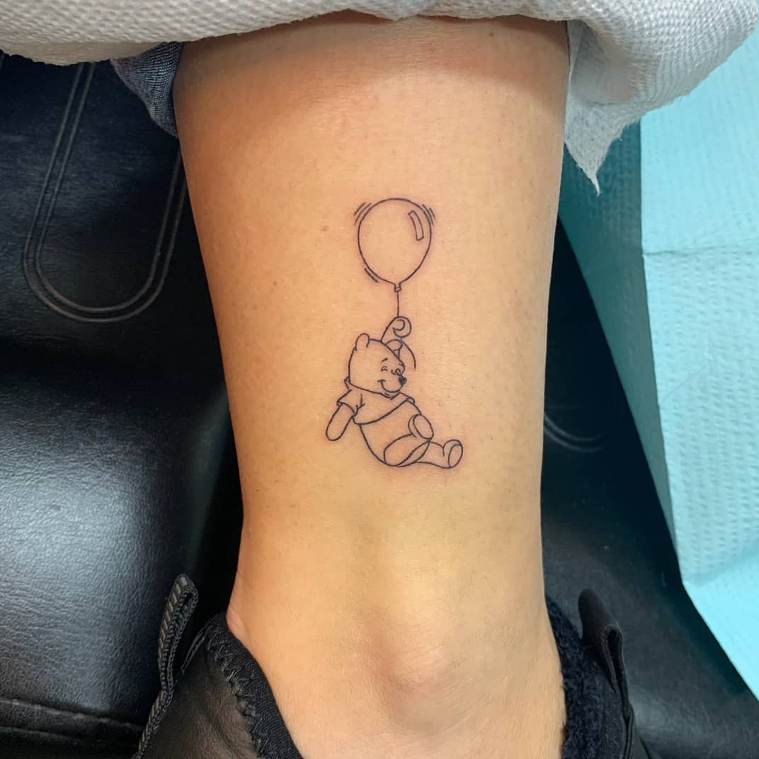 Outline tattoo of Winnie the Pooh holding onto a balloon on a person's leg